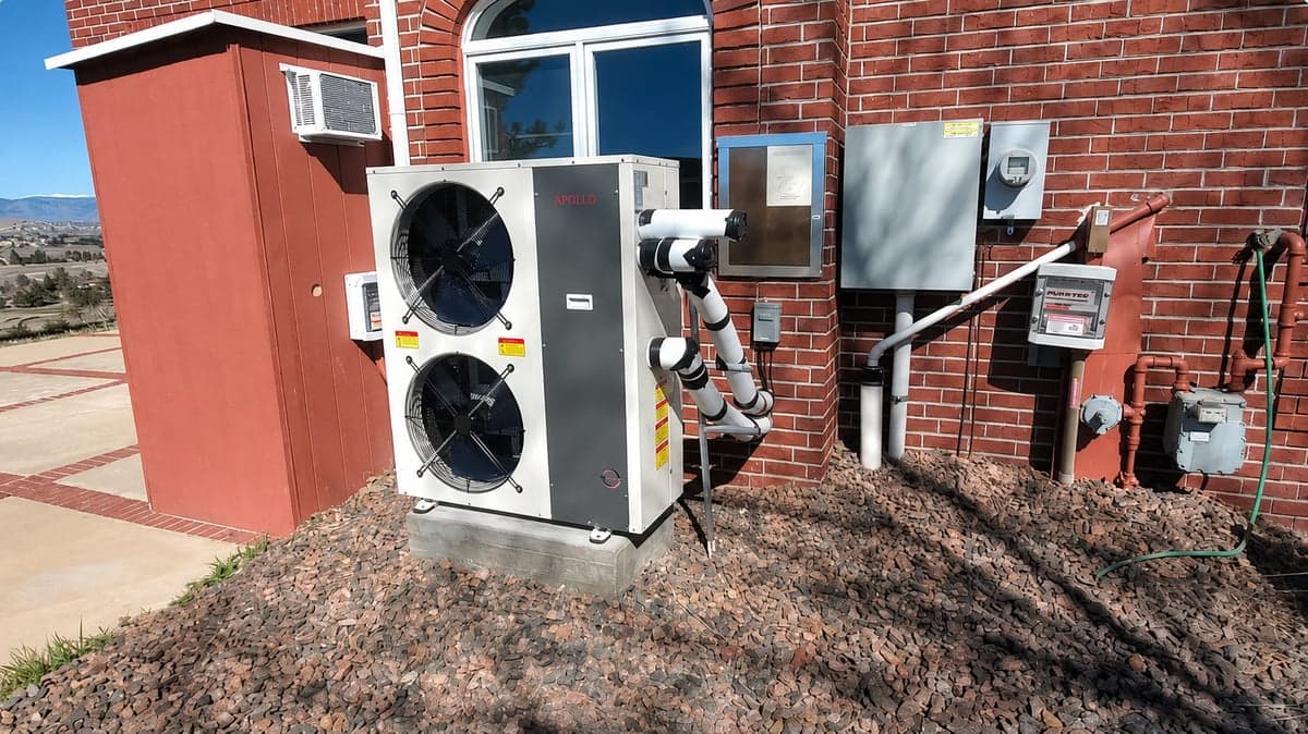 Apollo 6 ton air to water heat pump installed outdoors beside a brick home.