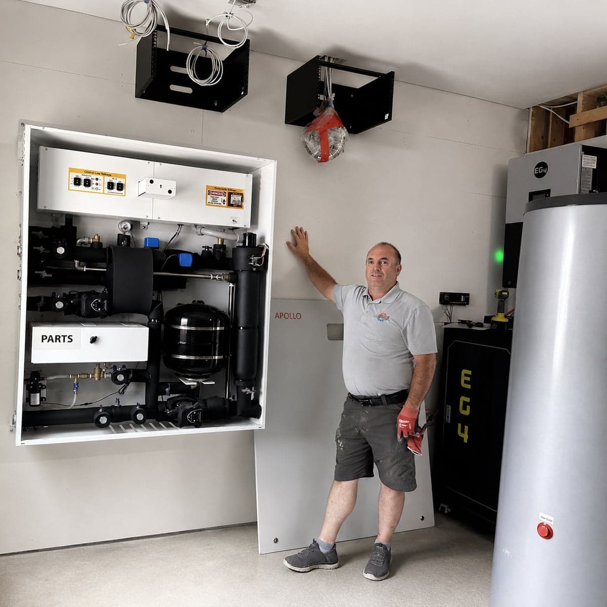 Installer standing beside an open Apollo hydro station with water tank and mechanical room equipment.