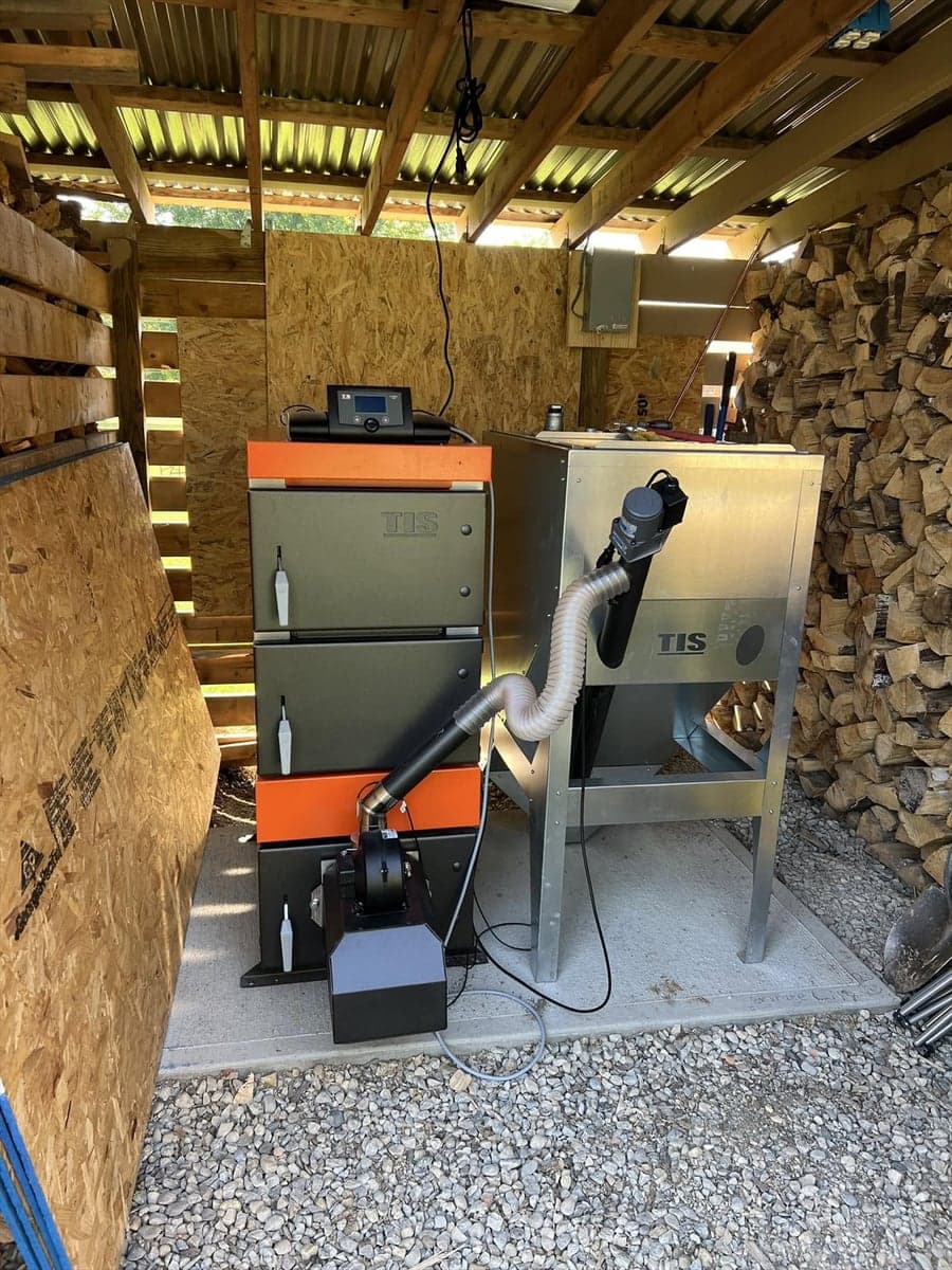Pellet Duo boiler and metal fuel hopper installed beside stacked firewood in a wood shed.