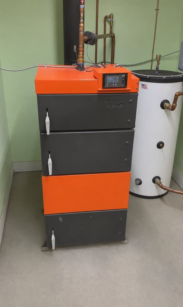 UNI wood boiler installed in a green mechanical room beside a vertical water storage tank.