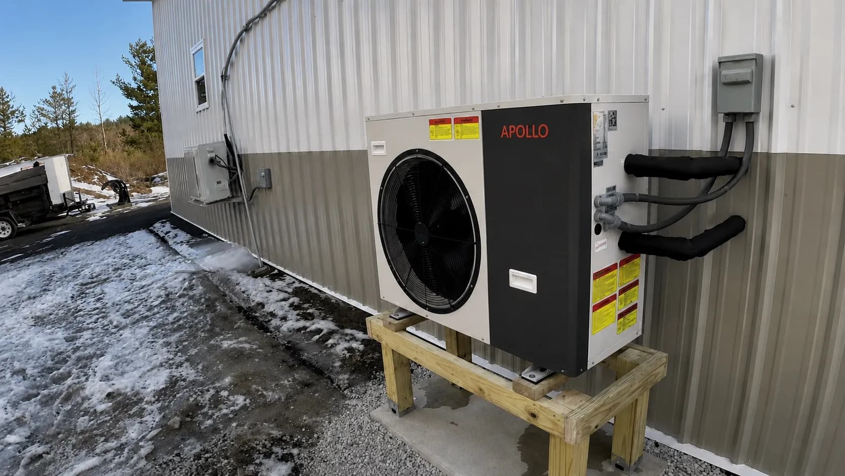 Apollo 3.5 ton air to water heat pump mounted outdoors on a wood stand beside a workshop.