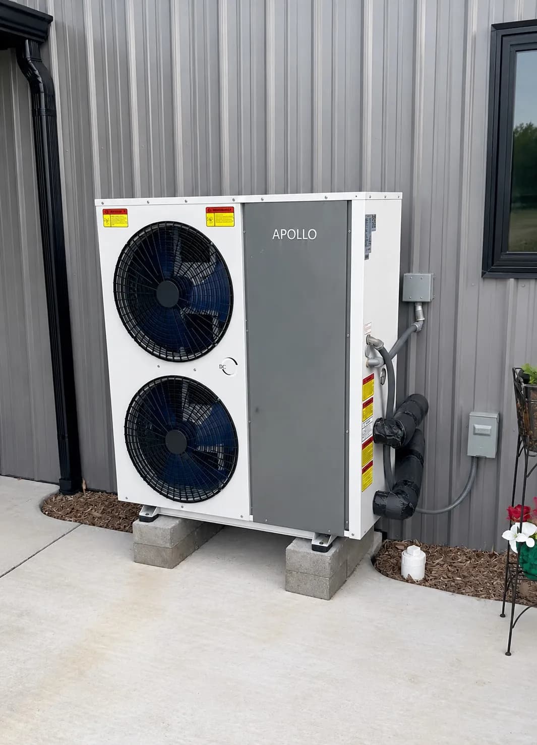 Apollo air to water heat pump installed outdoors on raised concrete blocks beside a metal-sided building.