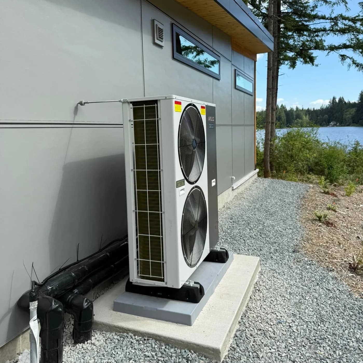 Dual-fan air-to-water heat pump installed beside a lakeside home on a concrete pad.
