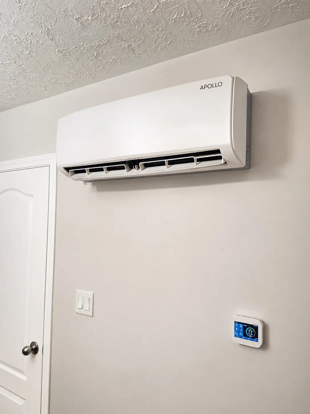 Apollo 1.5 ton wall fan coil installed above a thermostat with its air louvers open.