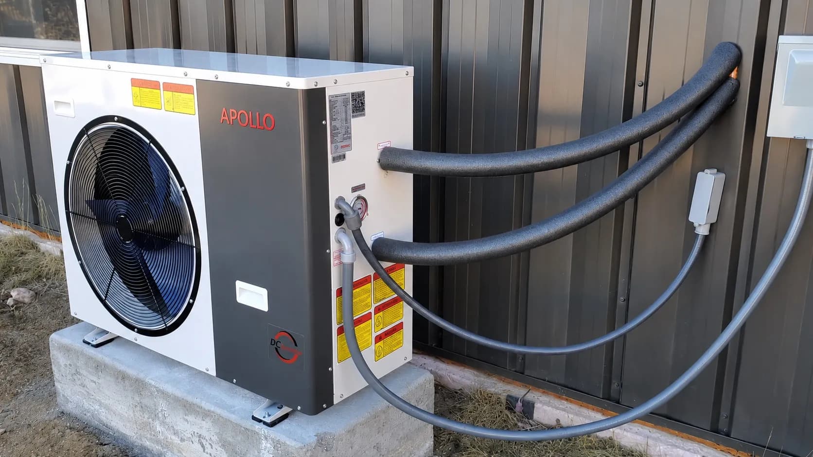 Apollo 3.5 ton air to water heat pump with insulated hydronic lines mounted beside a metal building.