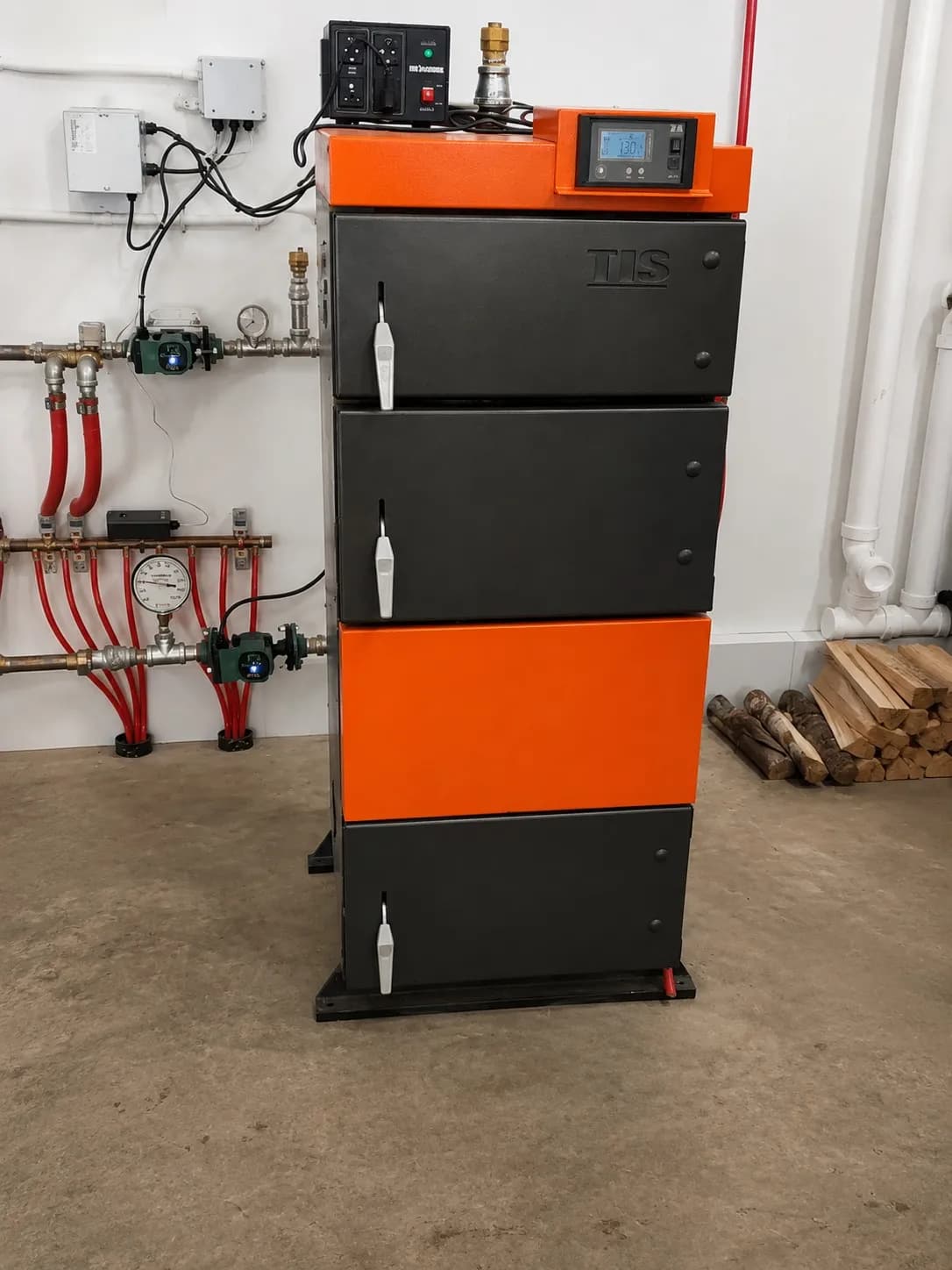 Black and orange boiler installed in a mechanical room with wall-mounted hydronic manifold, pumps, and control panel.