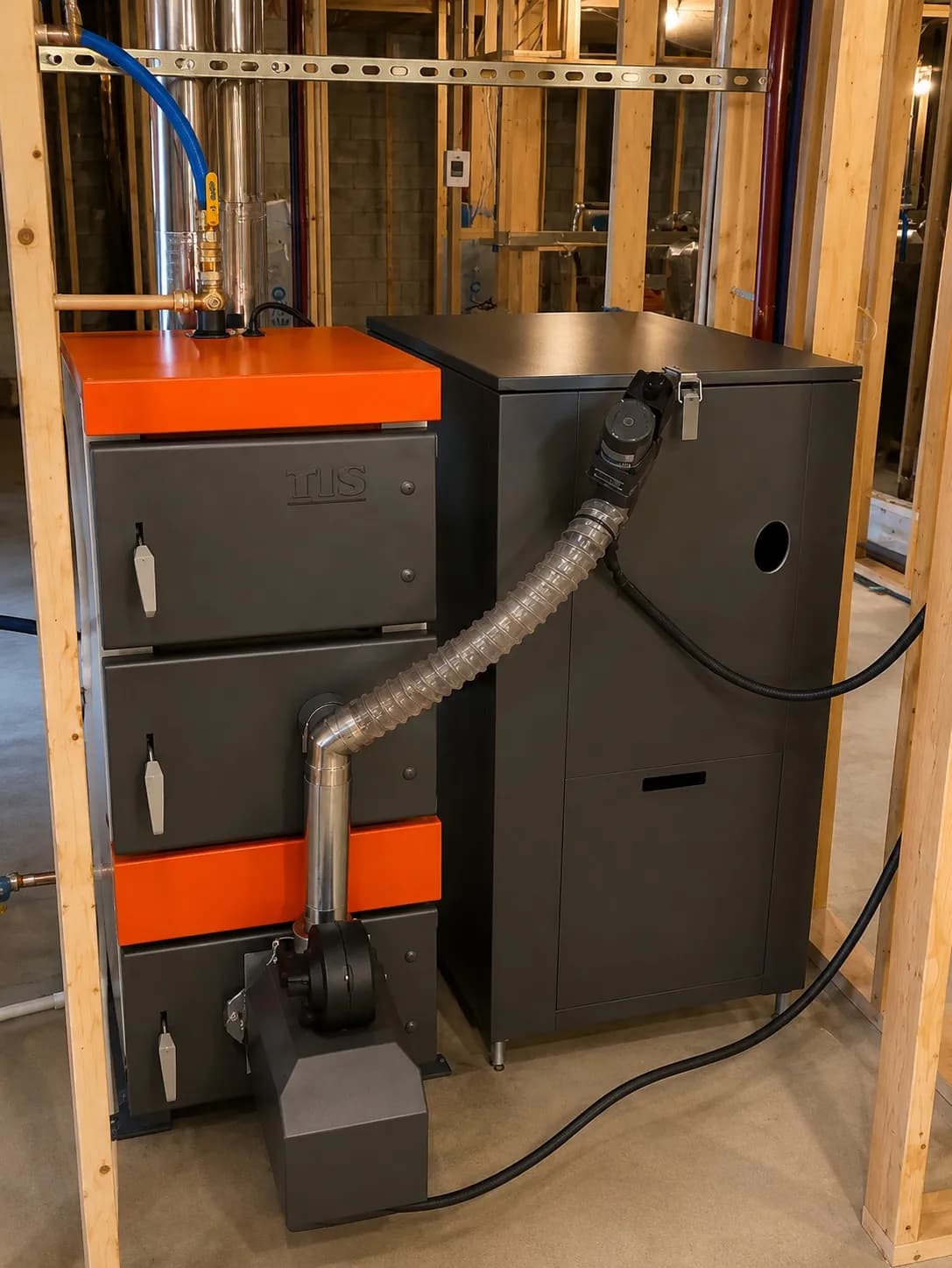 Black and orange pellet boiler connected to a black hopper and auger in an unfinished framed mechanical room.