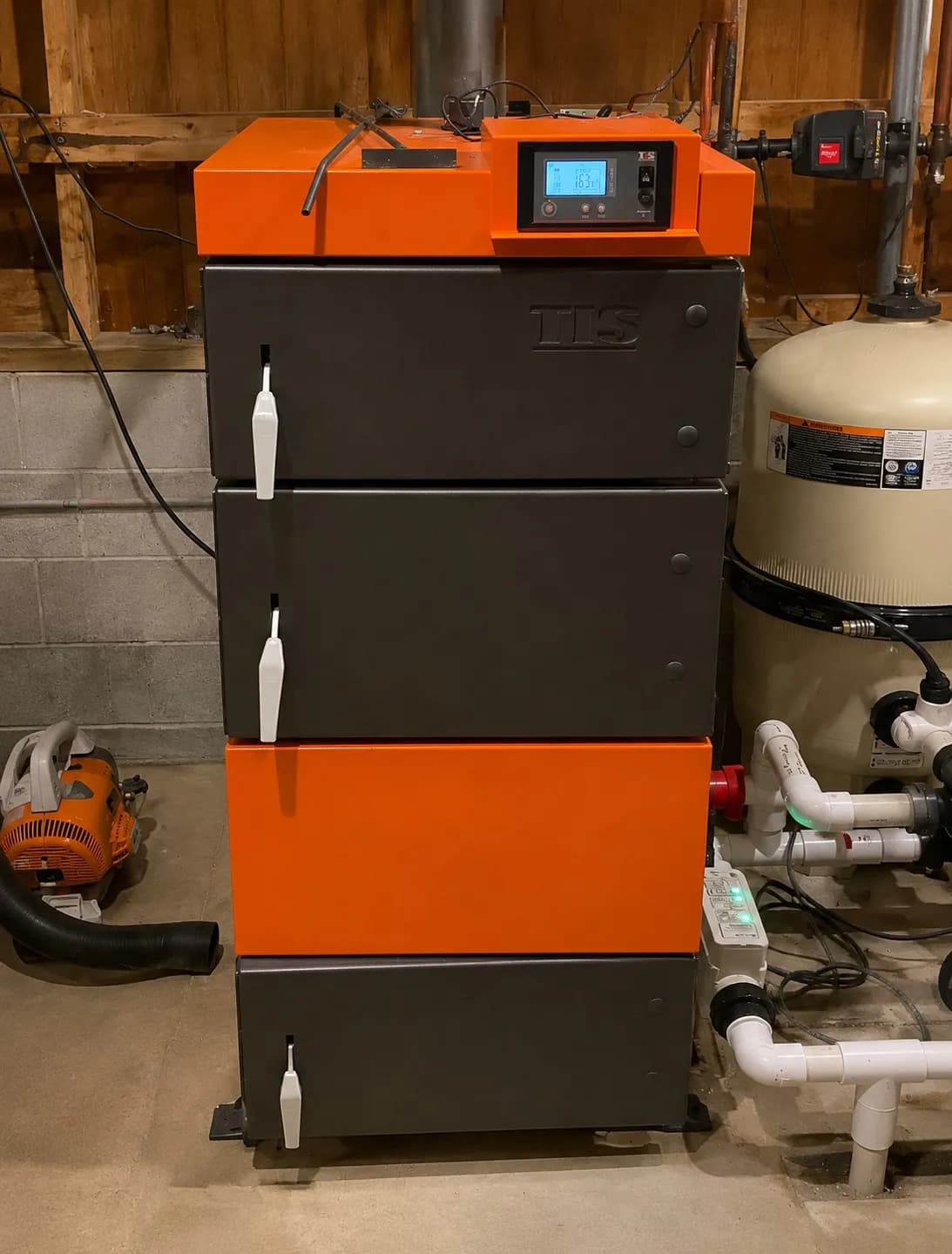 Black and orange boiler installed beside a water tank and hydronic piping in a basement mechanical room.