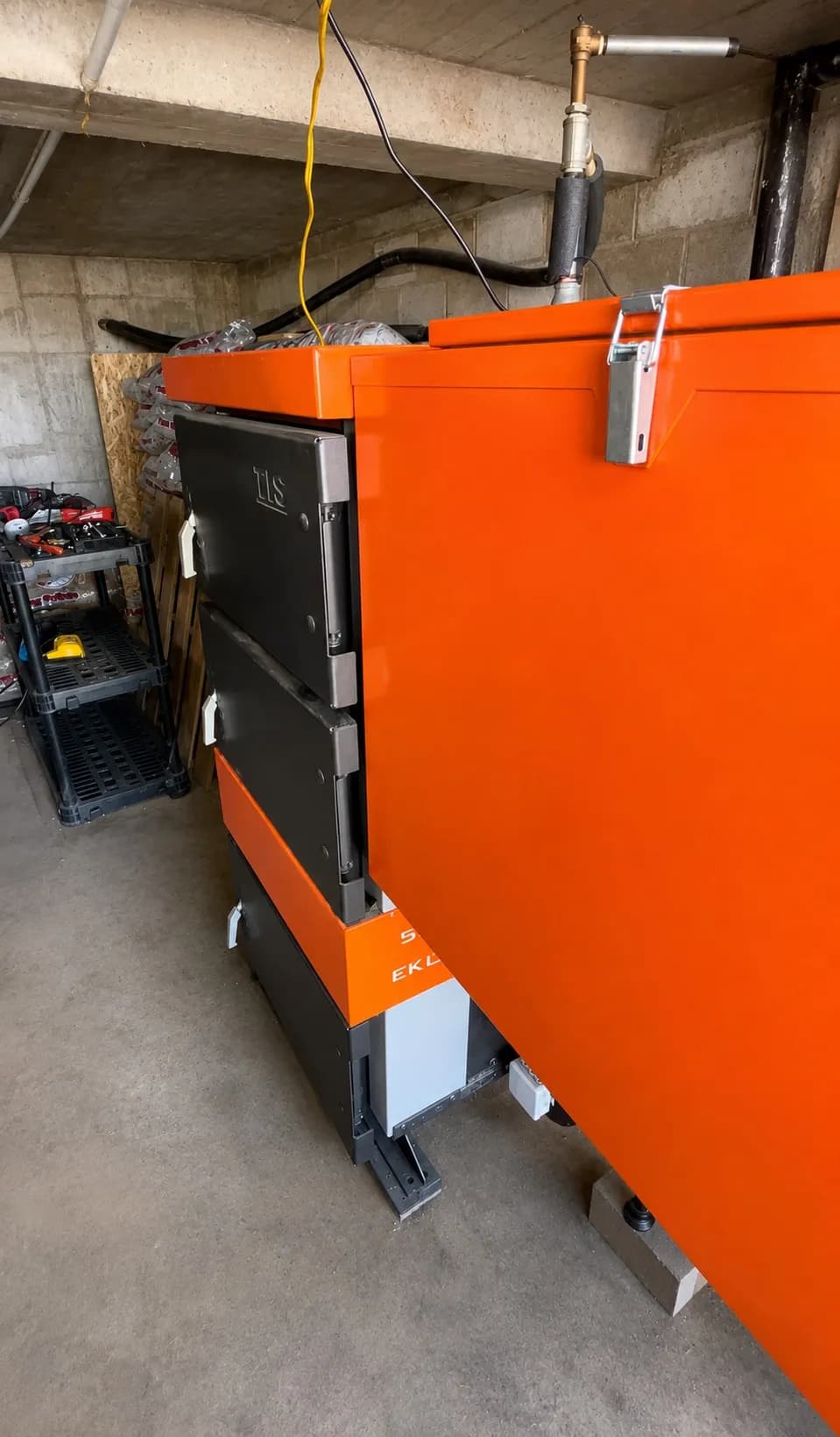 Side view of an orange EKO Duo hybrid coal boiler and integrated fuel hopper in a concrete mechanical room.